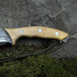 May include: Close-up of a handcrafted knife with a light wood handle and a textured metal blade. The handle is secured with three silver rivets. A green cord is attached to the handle. The knife rests on a stone surface.