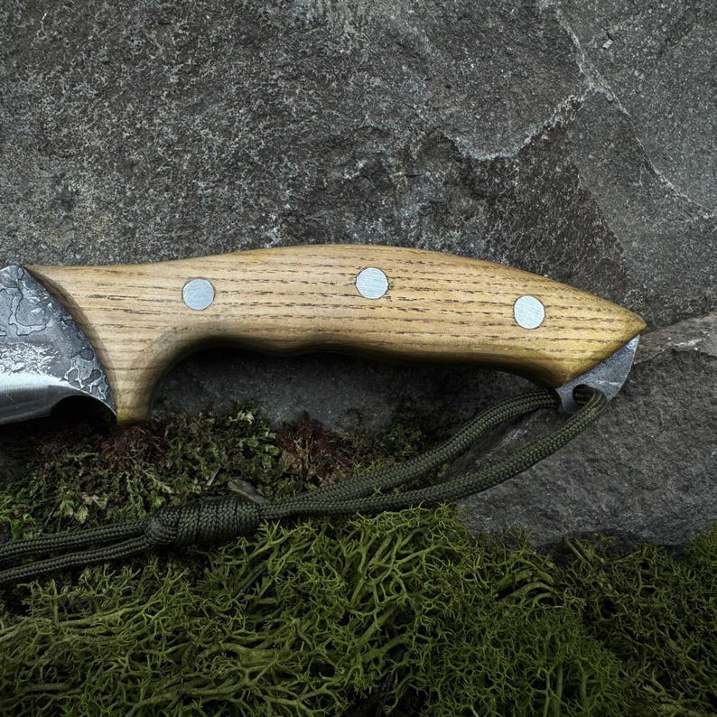 May include: Close-up of a handcrafted knife with a light wood handle and a textured metal blade. The handle is secured with three silver rivets. A green cord is attached to the handle. The knife rests on a stone surface.