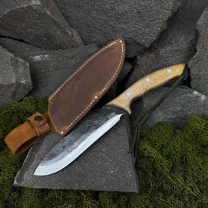 Puede incluir: Una funda de cuero marrón con un borde cosido y un cierre de presión metálico. Un cuchillo grande, negro, forjado a mano con mango de madera y un cordón de paracord verde oscuro está en la funda.