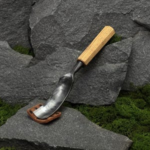 May include: A hand-forged metal spoon gouge featuring a curved, dark grey steel blade with a hammered texture and a cylindrical light brown wooden handle. The tool rests on a small, brown leather sheath with visible metal rivets at the base.