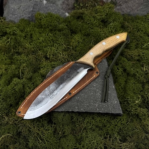 May include: A handcrafted knife with a light wood handle and a dark, textured blade, resting in a brown leather sheath. The knife is secured with rivets and a green cord. The background features green moss and a dark gray stone.