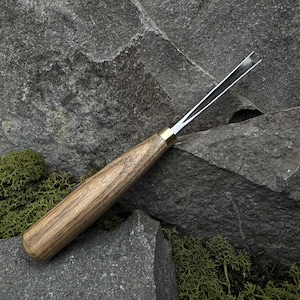 May include: A wood carving tool with a light brown wooden handle, brass ferrule, and a silver blade. The tool is designed for detailed woodworking and sits on a gray stone surface with green moss.