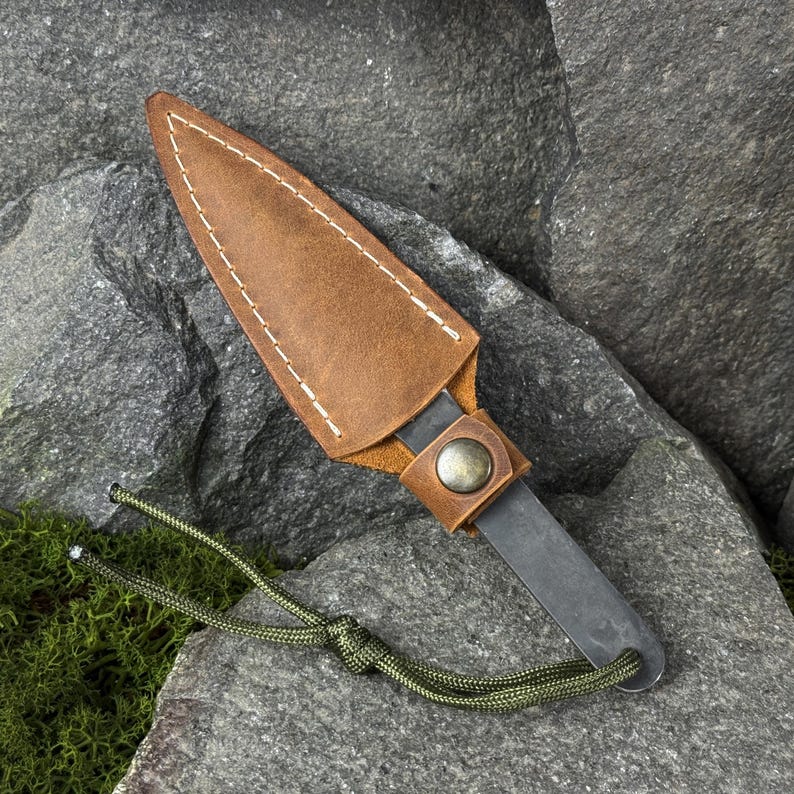May include: A small, dark metal throwing knife with a brown leather sheath. The sheath is stitched with white thread and has a snap closure. A green cord is attached to the knife's handle. The knife rests on a gray rock surface.