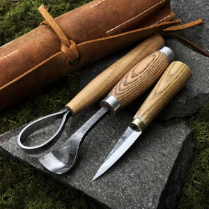 May include: A set of three wood carving tools with wooden handles and metal blades, alongside a rolled brown leather tool case. The tools are arranged on a dark gray stone surface, suggesting a craft or hobbyist theme.
