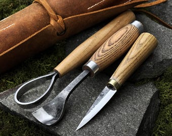Op de afbeelding: Een set van drie houtsnijgereedschappen met houten handvatten en metalen bladen, samen met een opgerolde bruine leren gereedschapskist. De gereedschappen zijn gerangschikt op een donkergrijze stenen ondergrond, wat duidt op een ambachtelijk thema.