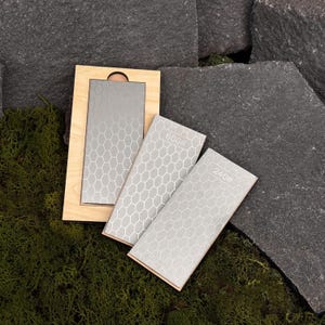 Pode incluir: Um conjunto de tr&ecirc;s pedras de amolar cinzentas com um padr&atilde;o de favo de mel, rotuladas 240# e 600#, e uma base de madeira. As pedras s&atilde;o projetadas para afiar facas e ferramentas. O fundo apresenta musgo e pedra cinza escura.
