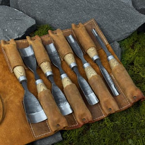 May include: A set of wood carving chisels with wooden handles and metal blades, stored in a brown leather tool roll. The chisels have a variety of blade shapes and sizes. The tool roll is open, displaying the tools against a backdrop of rocks and greenery.