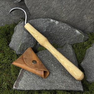 May include: A curved, silver-colored blade with a wooden handle and a small, brown leather pouch. The tool is designed for harvesting or gardening. The handle is made of light-colored wood. The pouch has a metal snap closure.