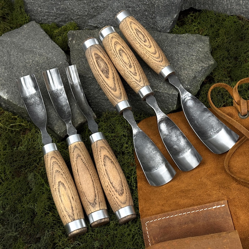 May include: A set of six wood carving gouges with curved metal blades and light brown wood handles featuring a prominent grain pattern. Each tool has a silver metal ferrule connecting the blade to the handle, arranged in a group.