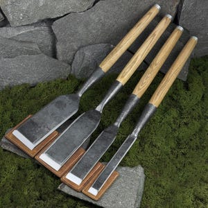 Puede incluir: Cuatro formones con mangos de madera y hojas de metal se exhiben sobre musgo verde. Cada formón descansa sobre una pequeña funda cuadrada de cuero con un inserto protector blanco. Los mangos tienen un patrón de veta de madera clara.