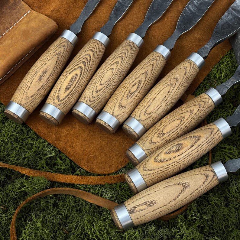 May include: A set of wood carving tools with light brown wooden handles and silver metal accents. The blades are dark gray. The tools are arranged on a brown leather surface, with a leather pouch visible in the background. The wood grain is visible.