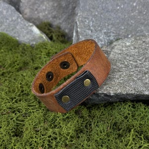 May include: A brown leather cuff bracelet with black elastic and brass-colored rivets. The bracelet has three black snaps for closure and two brass-colored rivets on the elastic section. The cuff is resting on a bed of green moss.