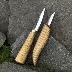 May include: Two wood-handled knives with sharp, silver blades. One knife has a brass collar. The handles have a light brown wood grain pattern. The knives are resting on a gray stone surface with green moss.