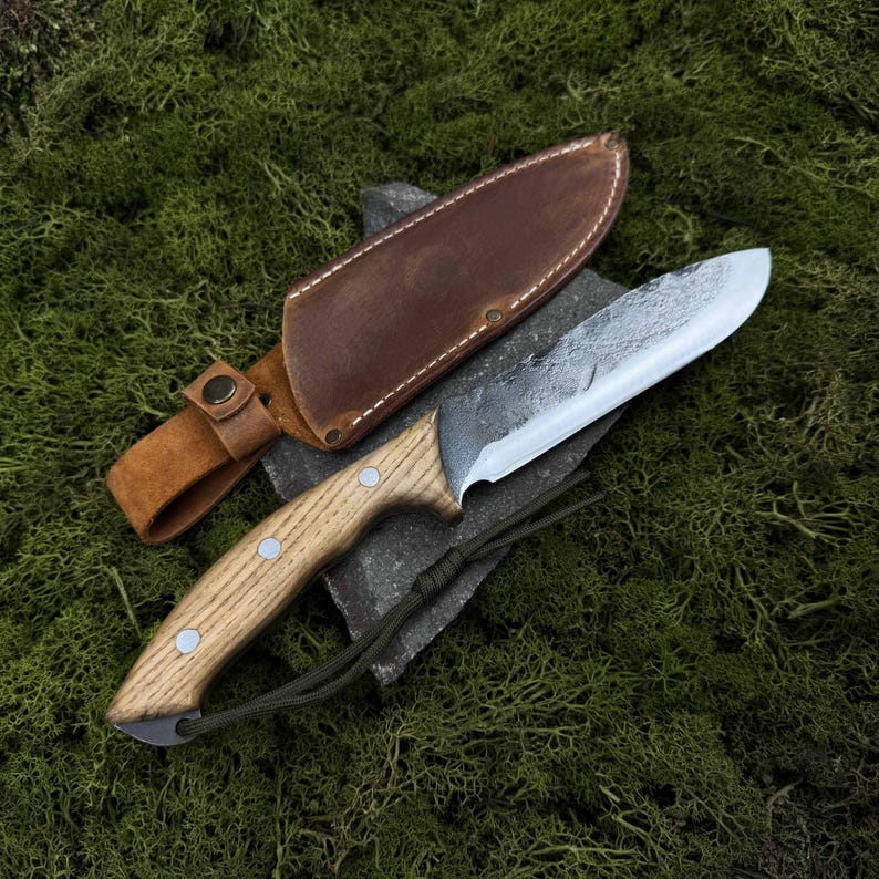 May include: A handcrafted knife with a light wood handle and a silver blade, resting on a dark stone. The knife is accompanied by a brown leather sheath. The handle has three silver rivets. A green cord is attached to the knife.
