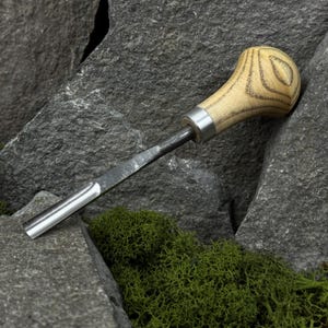 May include: A wood carving chisel with a light-colored, rounded wooden handle featuring a circular grain pattern. The metal blade is silver and extends from the handle. The chisel rests on gray rocks with green moss.