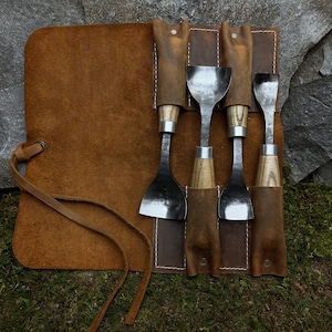 May include: A set of five wood carving tools with wooden handles and metal blades are rolled up in a brown leather tool roll with a strap.