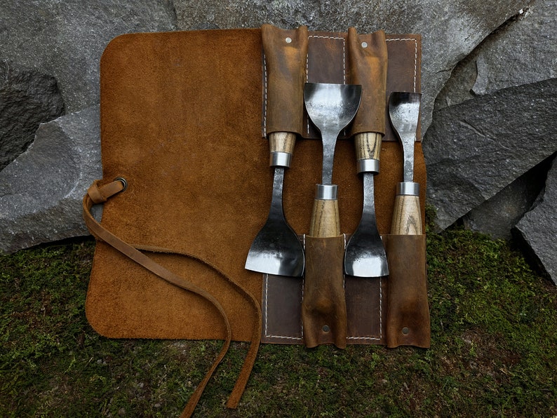 May include: A set of five wood carving tools with wooden handles and metal blades are rolled up in a brown leather tool roll with a strap.