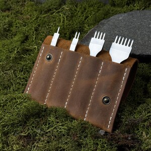 Może przedstawiać: Brązowy skórzany uchwyt na narzędzia z białymi przeszyciami, mieszczący cztery metalowe narzędzia do obróbki skóry. Narzędzia mają różne kształty końcówek. Uchwyt jest zabezpieczony dwoma metalowymi zatrzaskami i leży na zielonym mchu.