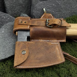 May include: Close-up of a brown leather axe sheath with white stitching and a metal buckle. The sheath covers the axe head and part of the handle. The axe head is dark metal, and the handle is light wood.