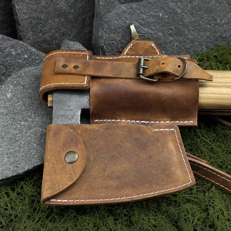 May include: Close-up of a brown leather axe sheath with white stitching and a metal buckle. The sheath covers the axe head and part of the handle. The axe head is dark metal, and the handle is light wood.