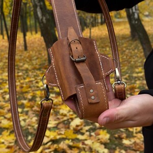 May include: A brown leather holster with a shoulder strap. The holster has a buckle closure and white stitching. The strap is attached with metal clasps. The background is a blurred view of a forest with yellow leaves.