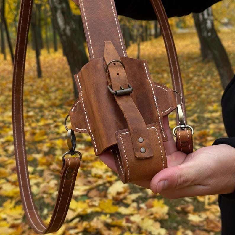May include: A brown leather holster with a shoulder strap. The holster has a buckle closure and white stitching. The strap is attached with metal clasps. The background is a blurred view of a forest with yellow leaves.