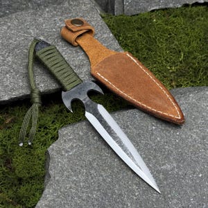 Puede incluir: Una daga artesanal con una afilada hoja plateada y un mango oscuro y texturizado envuelto en cordón verde. Presenta un diseño curvo único cerca del mango y viene con una funda de cuero marrón. La daga descansa sobre una superficie de piedra gris.