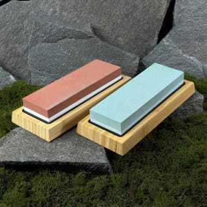 May include: Two sharpening stones, one red and one blue, sit on bamboo bases. Each stone has a white and black layer. The rectangular stones are displayed on green moss, with a backdrop of grey stone.