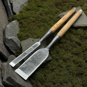 Puede incluir: Dos cinceles para carpintería con mangos de madera y hojas de metal. Un cincel es ancho y plano, el otro es estrecho. Las herramientas se colocan sobre un fondo de musgo y piedra.