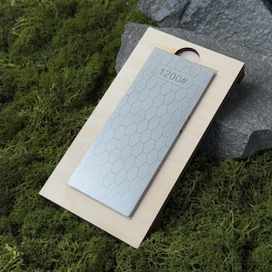 May include: A rectangular sharpening stone with a honeycomb pattern and the number "1200#" printed on it, set in a light-colored wooden base. The stone is gray and the base is light brown. The background is green moss.