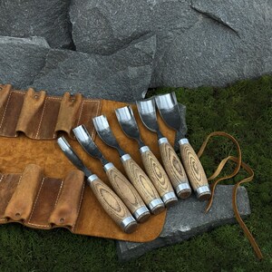 May include: A set of six wood carving gouges with curved metal blades and light brown wooden handles, arranged in a row on a tan leather tool roll with individual slots and leather tie strings, featuring visible wood grain on the handles.