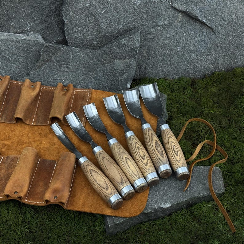 May include: A set of six wood carving gouges with curved metal blades and light brown wooden handles, arranged in a row on a tan leather tool roll with individual slots and leather tie strings, featuring visible wood grain on the handles.