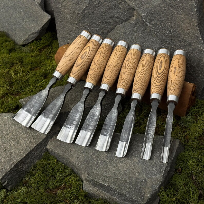 May include: A set of eight wood carving chisels with wooden handles and silver-colored blades. The handles are a light brown color with a wood grain pattern. The chisels are arranged on a gray stone surface.
