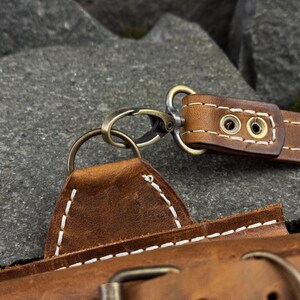 May include: Close-up of a brown leather strap with a metal clasp and ring. The leather is stitched with white thread. The metal hardware has a bronze finish. The strap appears to be part of a bag or accessory.
