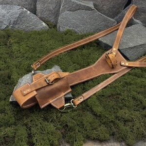 May include: A brown leather shoulder holster with a belt loop and adjustable straps. The holster features a small pouch and is secured with buckles and metal hardware. The stitching is white, and the leather has a distressed appearance.