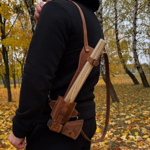 Könnte beinhalten: Eine braune Leder-Axtscheide mit Schulterriemen, die eine Holzaxt hält. Die Scheide hat einen Schnallenverschluss und eine kleine Tasche. Die Axt hat einen Holzgriff und eine Metallklinge.