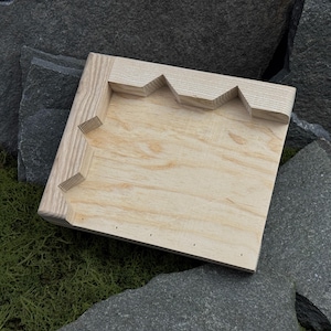 Könnte beinhalten: Ein helles Holztablett mit einem einzigartigen Zickzack-Kantendesign. Das Tablett ist quadratisch und weist ein natürliches Holzmaserungsmuster auf. Es ist vor einem Hintergrund aus grauen Steinen und grünem Moos platziert.