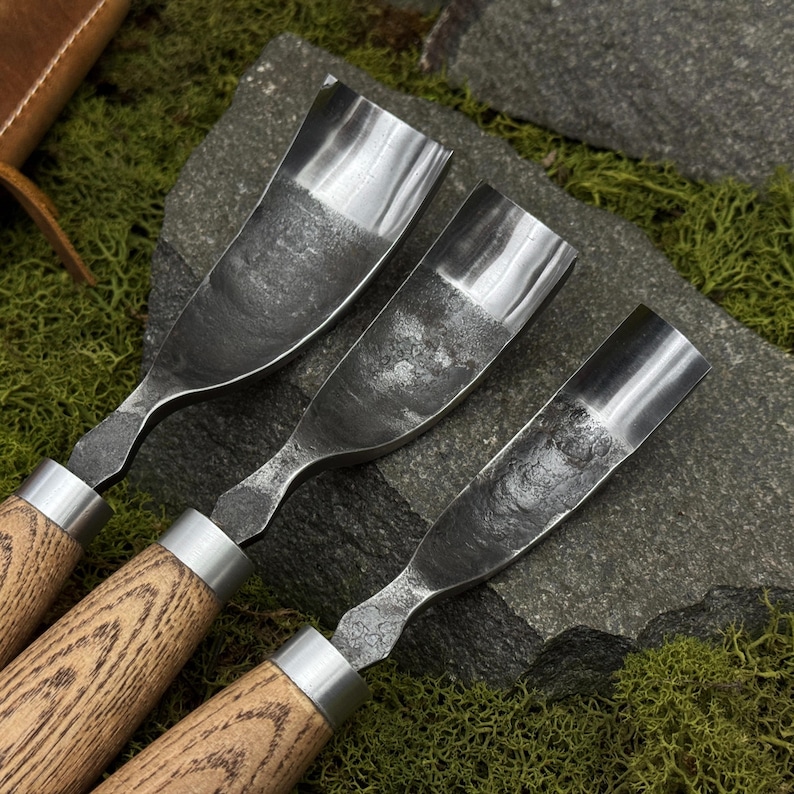 May include: Three wood carving gouges with curved, forged steel blades featuring a textured, dark finish and polished edges. Each tool has a light-colored wooden handle secured with a silver-toned metal ferrule, arranged in a descending size order.