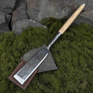 Puede incluir: Un cincel largo y plano con un mango de madera y una funda de cuero. El cincel está hecho de metal y tiene un borde afilado. El mango está hecho de madera y tiene un color marrón claro. La funda está hecha de cuero y tiene un color marrón oscuro.