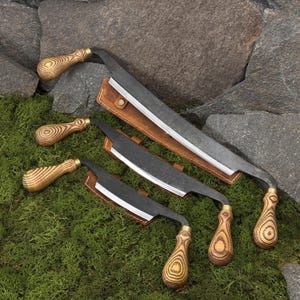 May include: A set of four curved, hand-forged knives with wooden handles and leather sheaths. The blades are dark gray, and the handles feature a light wood grain pattern. The knives are arranged on a bed of green moss against a backdrop of gray stone.