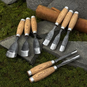 May include: A collection of wood carving chisels with wooden handles and metal blades. The chisels are arranged on a stone surface with a leather tool roll in the background. The handles have a light wood grain pattern and are accented with silver metal rings.