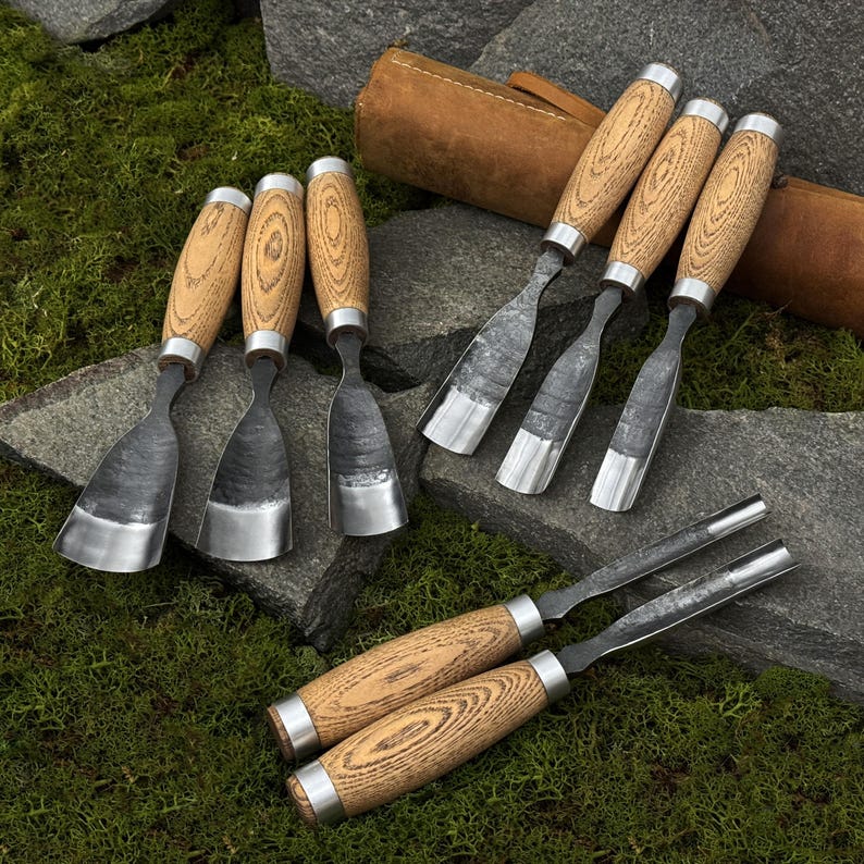 May include: A collection of wood carving chisels with wooden handles and metal blades. The chisels are arranged on a stone surface with a leather tool roll in the background. The handles have a light wood grain pattern and are accented with silver metal rings.