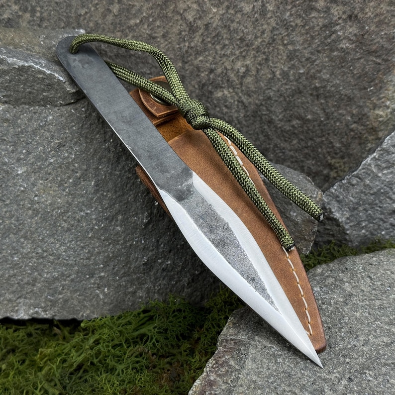 May include: A hand-forged throwing knife with a sharp, pointed blade and a textured, dark gray finish. It rests in a brown leather sheath with a green cord. The knife is designed for throwing and outdoor activities.