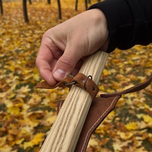 May include: A light-colored wooden staff with a brown leather strap and sheath. The staff is held by a hand, with the background showing fallen autumn leaves.