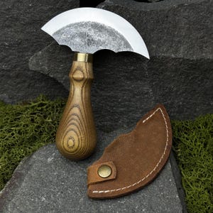 May include: A handcrafted, curved blade knife with a wooden handle and a leather sheath. The blade has a unique pattern, and the handle is made of light brown wood. The sheath is brown with white stitching and a brass button.