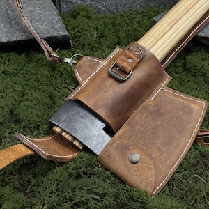 May include: A small axe with a wooden handle and a dark metal blade, partially encased in a brown leather sheath. The sheath has white stitching and a strap. The axe is resting on green moss.