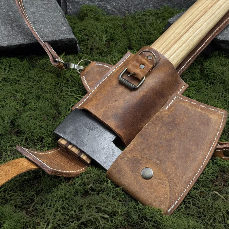 May include: A small axe with a wooden handle and a dark metal blade, partially encased in a brown leather sheath. The sheath has white stitching and a strap. The axe is resting on green moss.