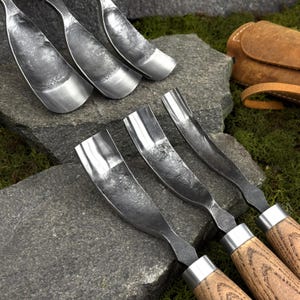 May include: A set of six hand-forged metal gouges with curved, textured steel blades and wooden handles. The tools feature a concave, scoop-like shape with a dark, hammered finish on the metal surfaces and light brown wood grain handles.