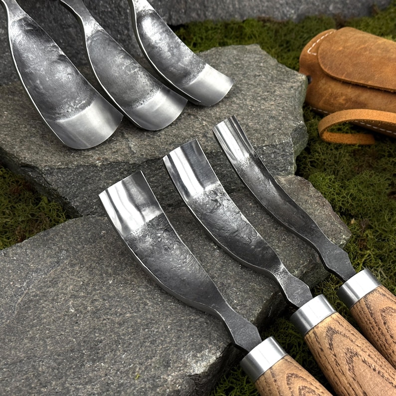 May include: A set of six hand-forged metal gouges with curved, textured steel blades and wooden handles. The tools feature a concave, scoop-like shape with a dark, hammered finish on the metal surfaces and light brown wood grain handles.