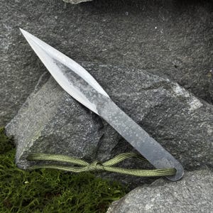May include: A hand-forged, silver-colored throwing knife with a sharp, pointed blade. The handle is a flat, textured metal with a small hole at the end. A green cord is attached to the handle. The knife rests on a gray rock surface.
